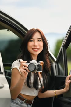 Charming millennial woman sitting in car with open door taking photos with vintage retro cameras during weekend road trip.