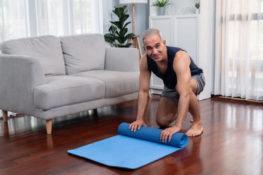 Active and sporty senior man preparing, rolling fitness exercising mat on living room floor at home. Home exercise as concept of healthy fit body lifestyle after retirement. Clout