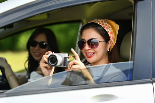 Smiling young woman taking photos from a car window with vintage retro cameras, enjoying road trip with her best friend.