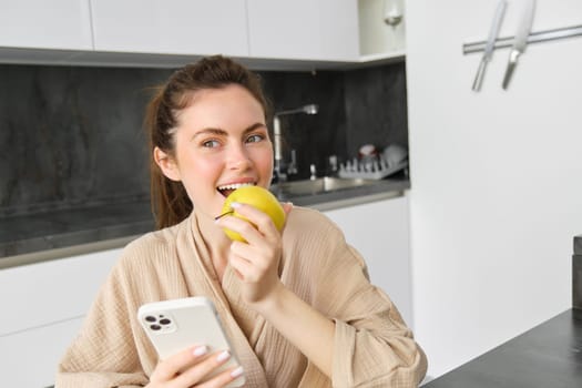 Portrait of happy, beautiful woman smiling, eating an apple in the kitchen, sitting at home in bathrobe, holding smartphone, using mobile phone.