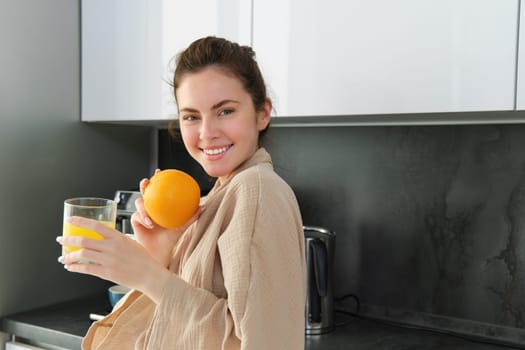 Portrait of happy woman in kitchen, wearing bathrobe, drinking orange juice, freshly squeezed drink, smiling and laughing, food and drink concept.