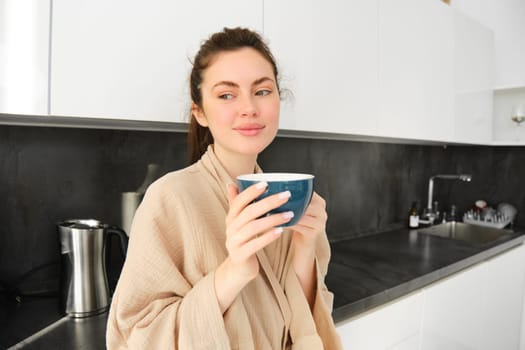 Romantic young woman drinking coffee in kitchen. Girl with mug in hands standing at home wearing a bathrobe, smiling with thoughtful face.