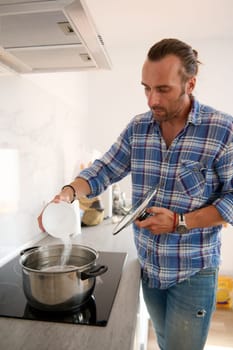 Caucasian young adult handsome male chef, a man 40 years old adding salt into a boiling water, in a saucepan standing on the black surface of an electric stove in modern white interior of home kitchen