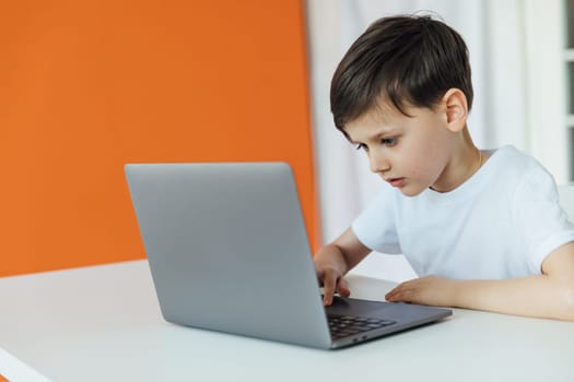 a boy sitting at a table playing at a laptop in a room against an orange wall