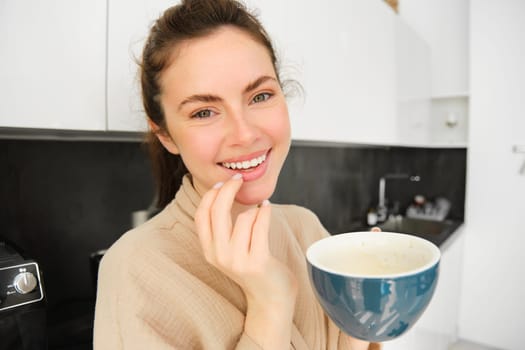 Close up portrait of attractive girl drinking coffee, holding cup with morning cappuccino and smiling, having a mug of delicious drink in the kitchen, wearing comfortable bathrobe.