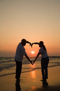 Mid-adult couple making heart shape with arms on beach at sunset.