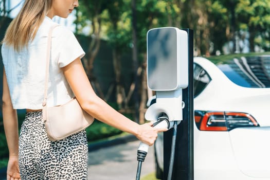 Young woman travel with EV electric car charging in green sustainable city outdoor garden in summer. Urban sustainability lifestyle by green clean rechargeable energy of electric BEV vehicle innards