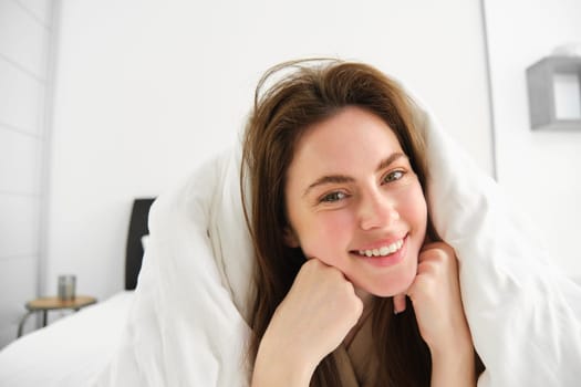 Portrait of attractive smiling woman relaxing in bed, lying covered in white sheets and warm duvet in morning, looking happy at camera.