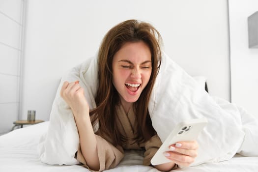 Cheerful brunette woman triumphing, looking at smartphone while lying in bed and celebrating, reading great news, winning, relaxing in her bedroom in morning.