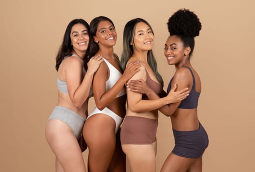 Four diverse young ladies confidently posing in their underwear, celebrating natural beauty and unity on beige studio background, looking and smiling at camera