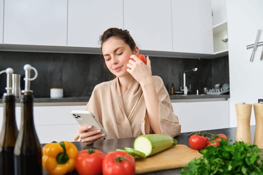 Young woman orders groceries on mobile app. Girl in bathrobe sits in the kitchen with vegetables, looking for recipe to cook dinner, using smartphone application.