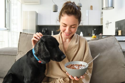 Happy young woman in bathrobe, enjoys spending time with her dog at home, eating cereals, having breakfast with puppy, sitting on sofa.