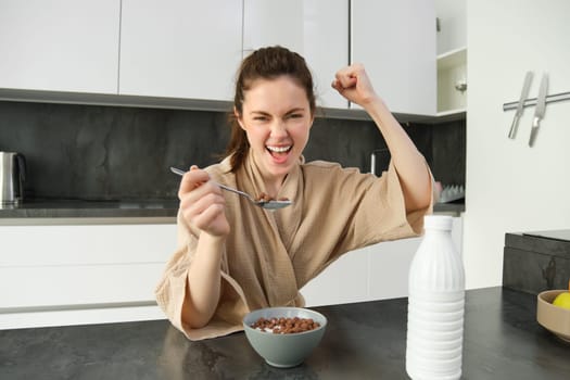 Portrait of enthusiastic young woman eating cereals with milk, looking excited and happy, sitting near kitchen worktop and having breakfast, raising hand up in triumph.