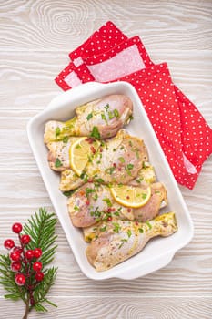Preparing festive Christmas Eve meal with marinated chicken drumsticks. Raw chicken legs with herbs, lemon in white ceramic casserole top view on light wooden kitchen table with Christmas decoration.
