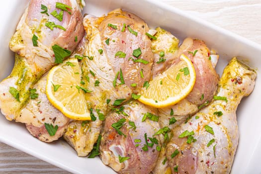 Raw uncooked chicken legs marinated with seasonings, herbs, lemon in white ceramic casserole top view on light wooden rustic background. Preparing healthy meal with marinated chicken drumsticks.