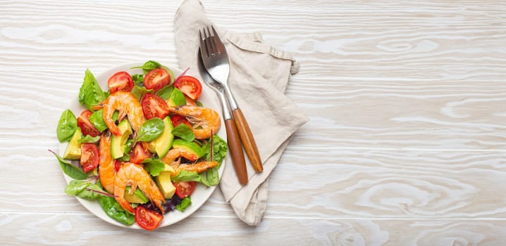 Healthy salad with grilled shrimps, avocado, cherry tomatoes and green leaves on white plate with cutlery on white wooden rustic background top view. Clean eating, nutrition and dieting, copy space..