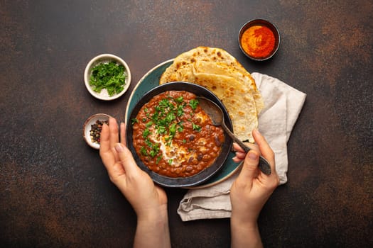 Female hands holding a bowl and eating traditional Indian Punjabi dish Dal makhani with lentils and beans served with naan flat bread, fresh cilantro on brown concrete rustic table top view.