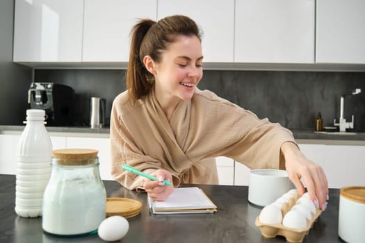 Attractive young cheerful girl baking at the kitchen, making dough, holding recipe book, having ideas.