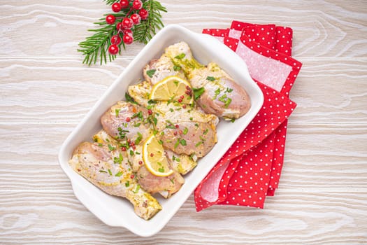 Preparing festive Christmas Eve meal with marinated chicken drumsticks. Raw chicken legs with herbs, lemon in white ceramic casserole top view on light wooden kitchen table with Christmas decoration.