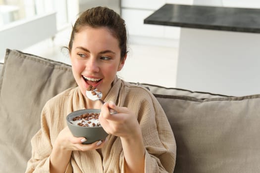 Portrait of woman on sofa in bathrobe, eating cereals with milk, watching tv and having breakfast.