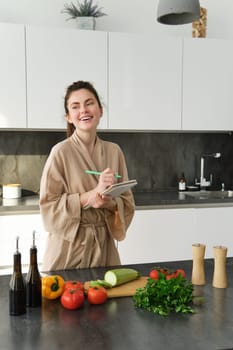 Vertical shot of young woman writing down food ideas, creating new meal, writing down grocery list or recipe in the notebook, standing in the kitchen near chopping board with fresh vegetables.