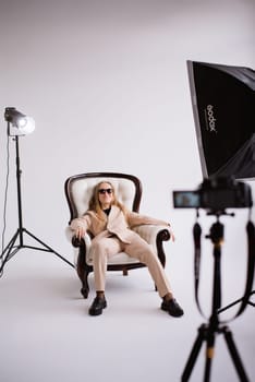 Female blogger, blonde in sunglasses sitting on chair in white photo studio, isolated. Dressed in a formal beige trouser suit