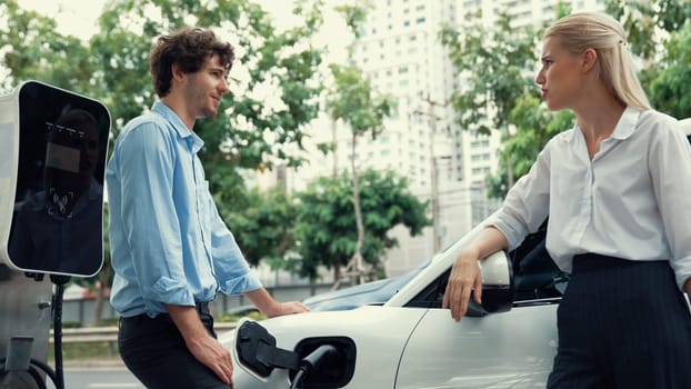 Progressive businessman and businesswoman install charger plug from charging station to electric car before driving around city center. Eco friendly rechargeable car powered by clean energy.