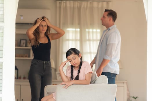 Annoyed and unhappy young girl sitting on sofa trapped in middle of tension by her parent argument in living room. Unhealthy domestic lifestyle and traumatic childhood develop to depression.Synchronos