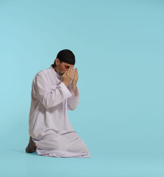 devout Muslim man kneels in pray, hands covering face in act of Namaz, fundamental Islamic ritual. Isolated against blue background. Faith, spirituality, and devotion in practice of Islamic prayer. High quality photo