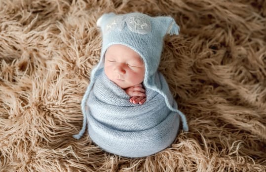 Adorable newborn baby boy swaddled in knitted blanket and wearing hat on fur studio portrait. Cute infant child kid sleeping