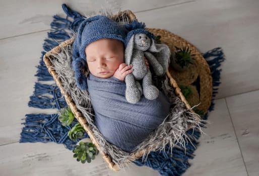 Adorable newborn baby boy swaddled in blanket holding knitted bunny toy and sleeping in basket studio portrait. Cute infant child kid napping