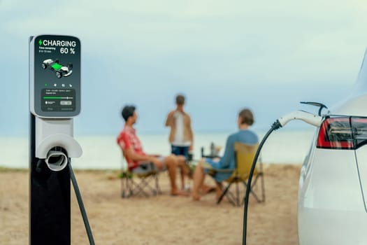 Alternative family vacation trip traveling by the beach with electric car recharging battery from EV charging station with blurred family enjoying the seascape background. Perpetual