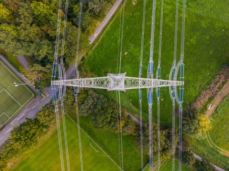 High voltage pylon with power lines between nature and sports field photographed directly from above with drone