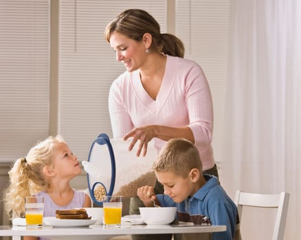 A beautiful family eating breakfast together. The mother is smiling and is pouring cereal for the daughter. Horizontally framed shot.