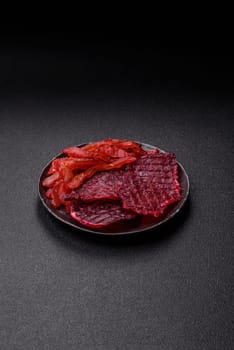 Dried beef slices, jerky, with cherry or plum flavor, salt and spices on a dark concrete background