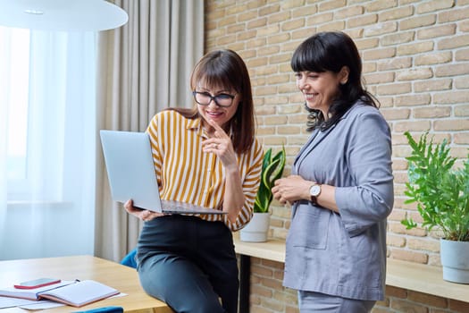 Two mature business women talking in modern office. Laughing communicating female colleague, 40s 50s age looking at laptop screen. Business ceo work law finance, mentoring consulting teamwork job