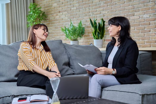 Two mature business women are talking in a modern office. Laughing communicating female colleague, 40s 50s age. Business, ceo, work, law, finance, mentoring, consulting, teamwork, job