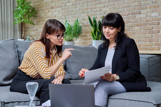 Two mature business women having conversation sitting on couch in office, discussing work, commercial projects. Communicating female colleague, 40s 50s age. Law finance mentoring consulting teamwork