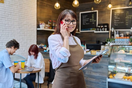 Portrait of confident middle aged woman coffee shop worker. Female in apron holding clipboard with orders looking at camera talking on cell phone with cafeteria customers. Small business work people