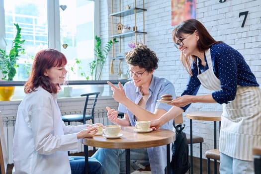 Woman in an apron with cup of coffee and plate of cake serving young people sitting at table in coffee shop, cafeteria, bakery. Food service occupation, small business, staff, job work, people concept