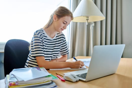 Teenage female student sitting at home at desk, writing in notebook using laptop computer. Girl studies online, remote lessons, prepares for tests, watches educational webinars. Technology adolescence