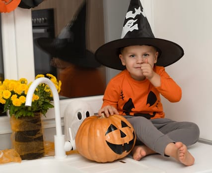 Halloween concept. A small, cheerful, handsome boy in a wizard's hat and an orange sweater sits on a table in the kitchen against the background of a pumpkin, a ghost and a bouquet of yellow flowers. Festive interior.