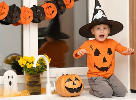 Halloween concept. A small, cheerful, handsome boy in a wizard's hat and an orange sweater sits on a table in the kitchen against the background of a pumpkin, a ghost and a bouquet of yellow flowers. Festive interior.