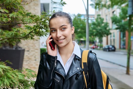 Young attractive teenage girl talking on mobile phone, outdoor on street of modern city. Teenager, college student with backpack, communicating on smartphone. Urban style, lifestyle, youth