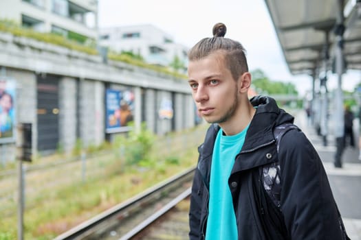 Young male waiting for electric train at city railway platform station. Guy serious student hipster 19, 20 years old with backpack. Urban transport, commuter train, passenger transportation, youth