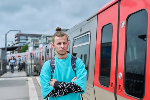 Young male waiting for electric train at city railway platform station. Guy serious student hipster 19, 20 years old with backpack. Urban transport, commuter train, passenger transportation, youth