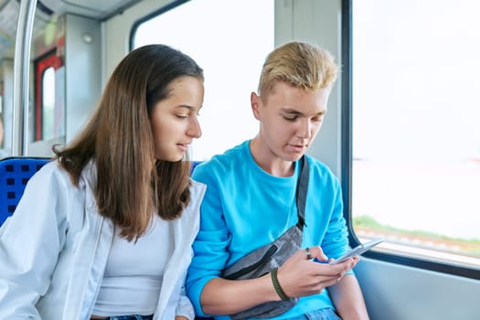 Teenage guy and girl commuter train passengers sitting together looking at smartphone screen. Adolescence, youth, lifestyle, friendship, communication, technology concept