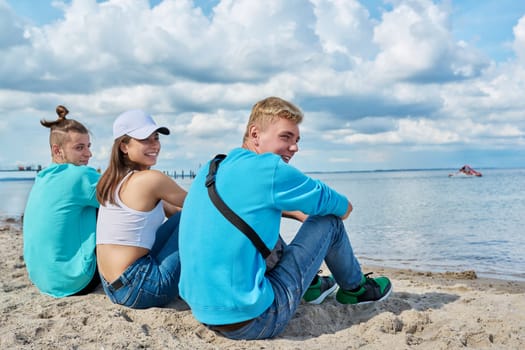 Back view of three teenage friends sitting on the sand at the beach, looking at sea, talking and laughing. Youth guys and girl, summer vacation, leisure, friendship, fun, lifestyle holiday concept