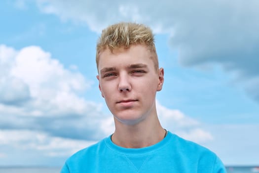 Headshot portrait of handsome smiling teenage guy, face of positive young male against blue sky in clouds. Youth, 18, 19, 20 years of age, lifestyle, enjoy fun, holiday vacation, outdoor