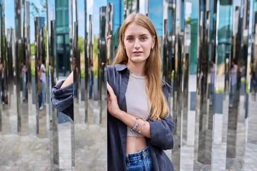 Portrait of young beautiful teenage female in modern city, fashionable smiling girl with long hair looking at camera, on mirror architecture. Beauty, fashion, youth, urban style concept